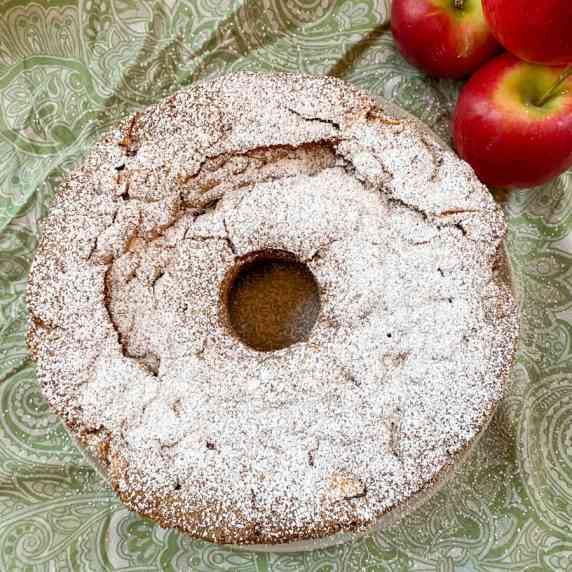 A round cake sprinkled with powdered sugar arranged on green paisley fabric with apples alongside.