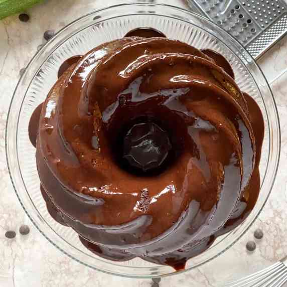 A chocolate bundt cake covered in chocolate sauce sits on a glass plate on a marble surface.