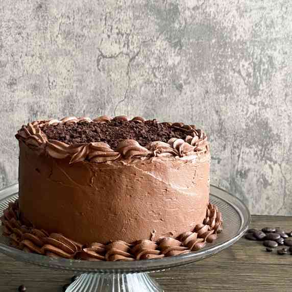 a chocolate layer cake sits on a glass cake stand on a wooden surface with a speckledgrey background