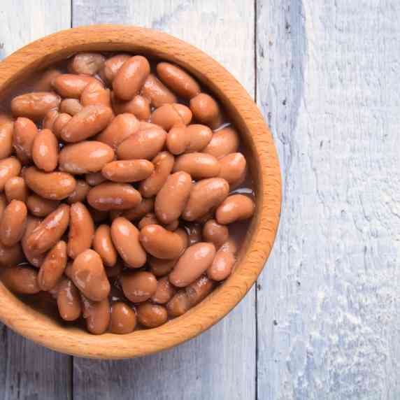 A wooden bowl of instant pot pinto beans rests on a light grey wooden table.