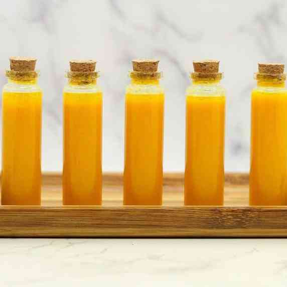 Orange colored Lemon Ginger Turmeric Wellness Juice Shots in tiny glass vials on a wooden plate.