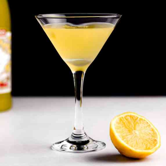 A limoncello martini on a white table with a black background, next to half an orange. 