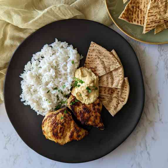 A plate of marinated chicken and rice served with pita bread and hummus