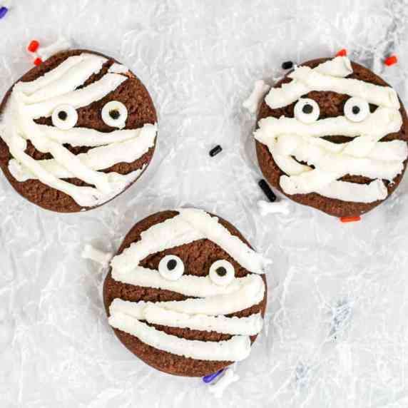 Three chocolate cookies close up decorated with frosting and candy eyes to look like mummies.