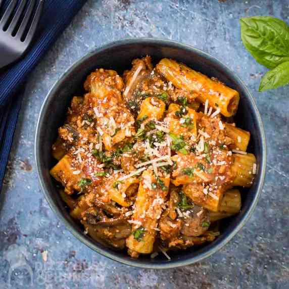 Side shot of rigatoni garnished with cheese and herbs in a blue bowl on a blue speckled surface.