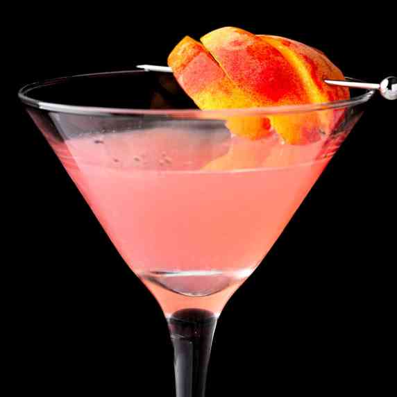 A pink peach cosmo cocktail garnished with peach slices on a black background.