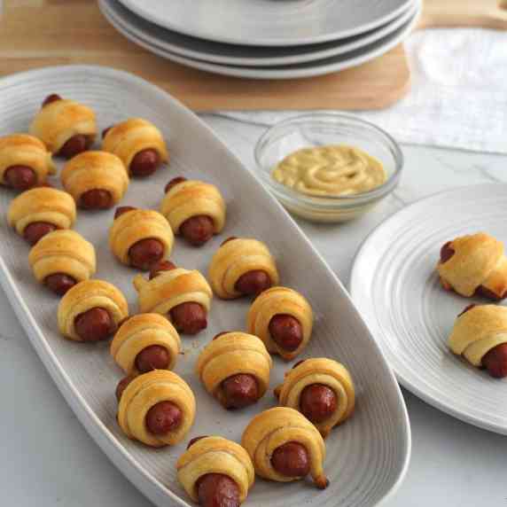 A serving plate of pigs in a blanket with a small dish of mustard and plates in the background.