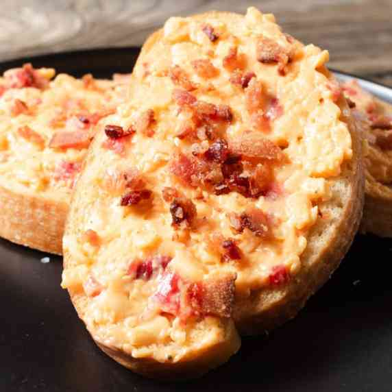 Pimento cheese and bacon topped crostini on a black cocktail plate.
