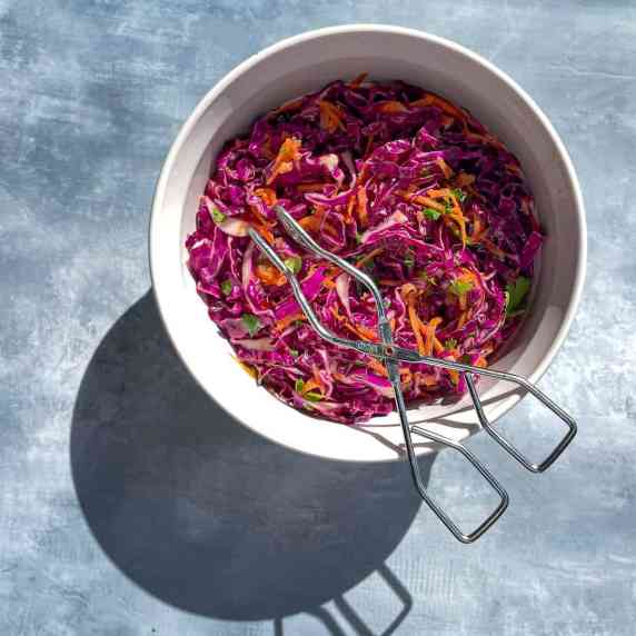 white bowl with shredded purple cabbage, carrots, cilantro, and tongs on blue background.