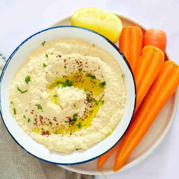 A small bowl filled with hummus drizzled with oil and chopped herbs. Some carrot sticks to the side.