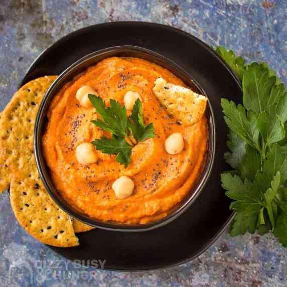 Overhead shot of red pepper hummus in a black bowl garnished with chickpeas, herbs and pepper. 