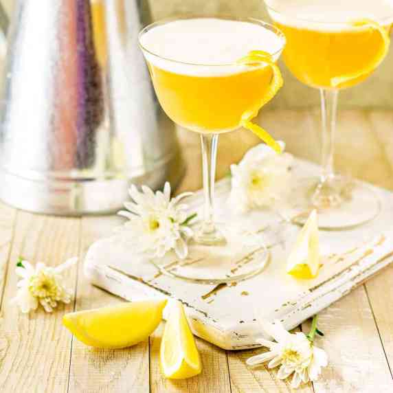 Two rum sour cocktails on a white wooden board with lemon slices and flowers around them.
