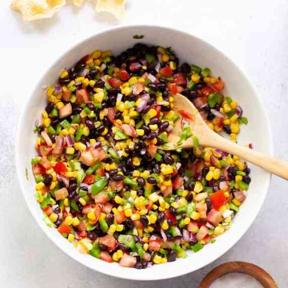 Overhead of a white bowl filled with fresh corn and black bean salsa with a wooden serving spoon.