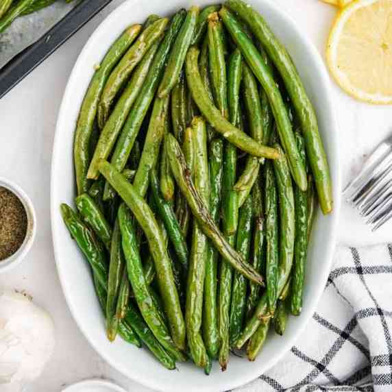 Overhead of an oval dish of oven roasted green beans with fresh lemon slices and forks nearby.