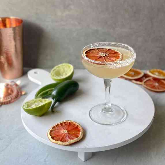 a skinny spicy margarita garnished with dried orange slice on a tray with jalapenos and limes.