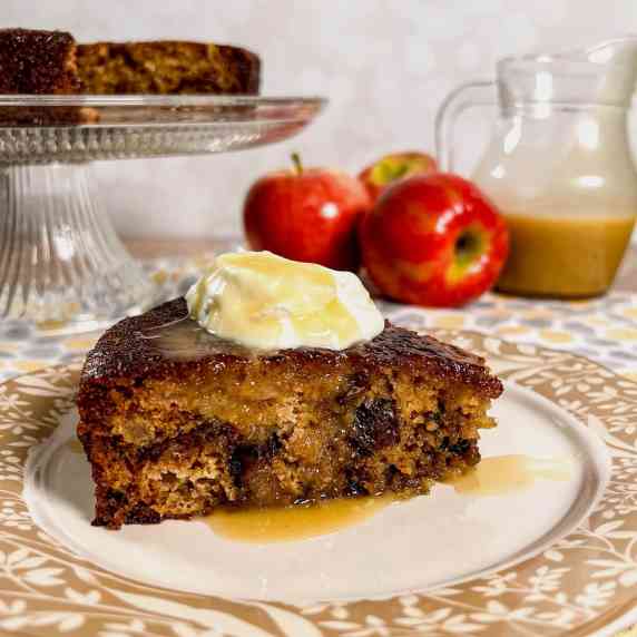 Slice of sticky toffee apple pudding on a plate with the rest of the cake on a cake stand.