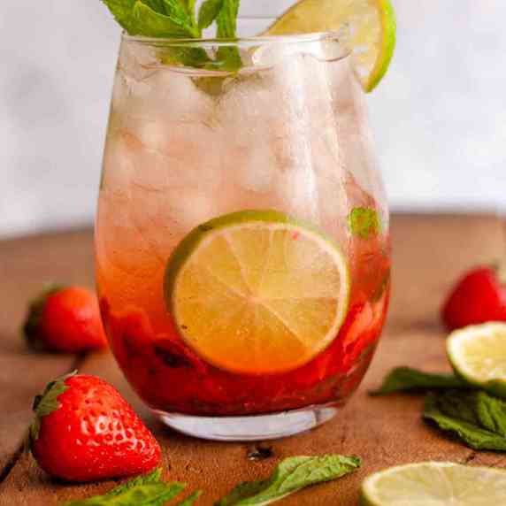 A close up of a strawberry mojito mocktail on a wooden surface next to lime slices and strawberries.