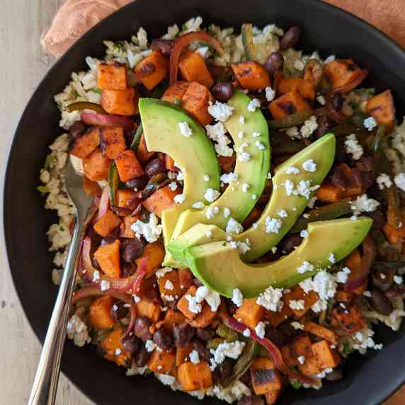 A grain bowl of brown rice, black beans, sweet potatoes, and veggies topped with avocado