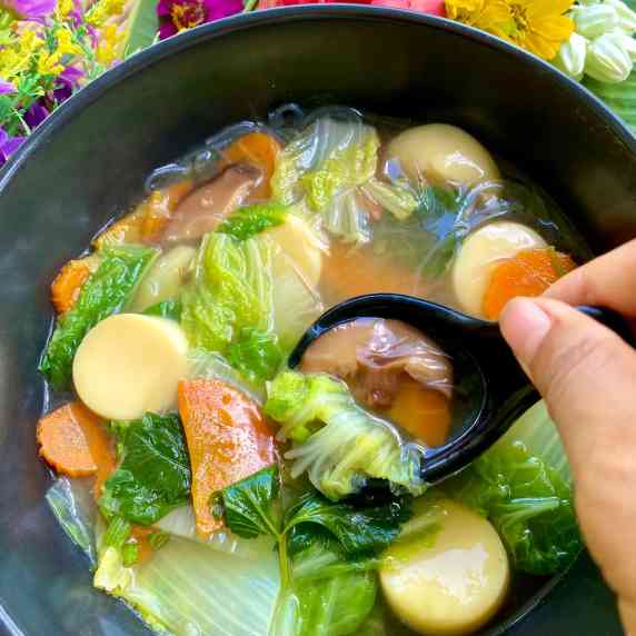 Hand holding black spoon over black bowl of Thai vegetable soup.