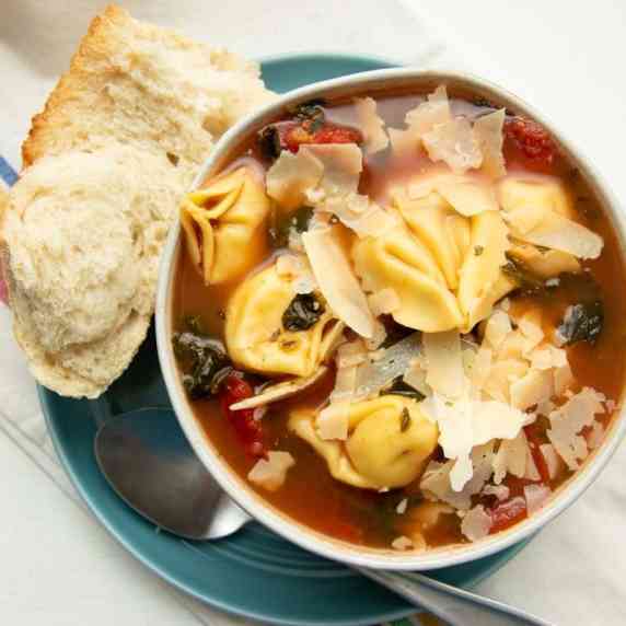A bowl of tortellini soup garnished with shaved parmesan rests on a plate with crusty bread.