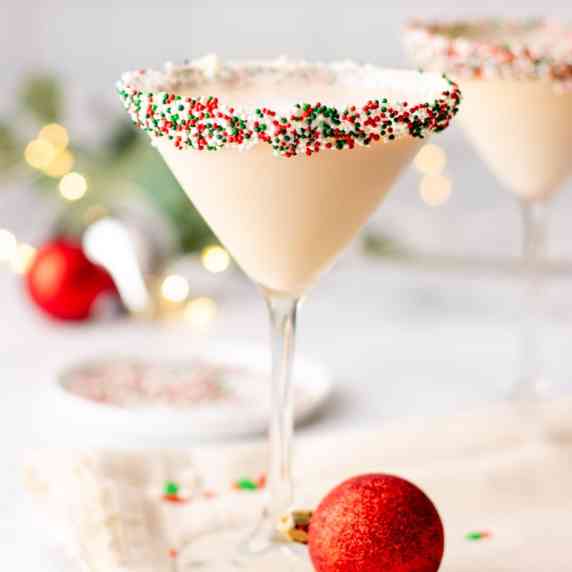 A creamy sugar cookie martini with a frosting and sprinkles coated rim stands with holiday ornaments