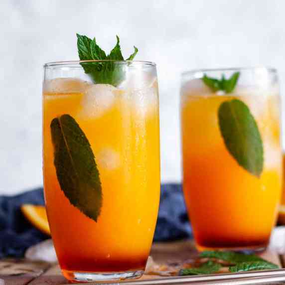 Two glasses of virgin sunrise mocktails on a wooden table next to orange wedges and mint leaves.