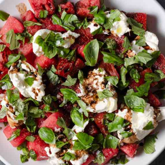 A plate of watermelon and burrata salad with basil, balsamic glaze, olive oil, and salt.