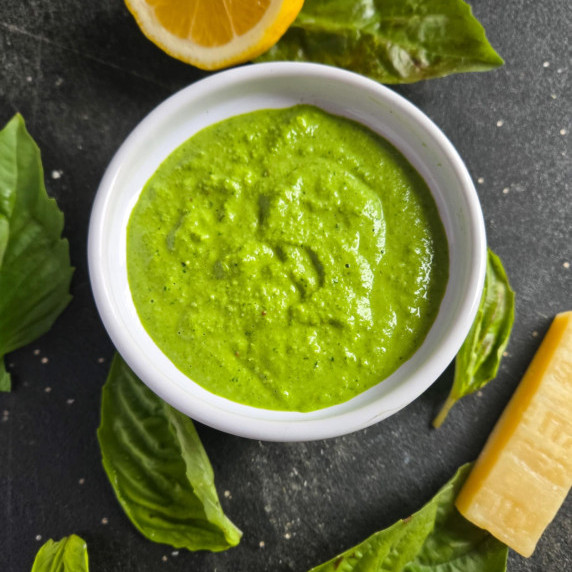 A white bowl filled with gorgeously green pesto surrounded by green basil, lemon, and hard cheese