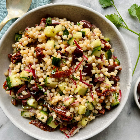 Salad with couscous, cucumbers, onion, sun dried tomatoes and olives in a white bowl.