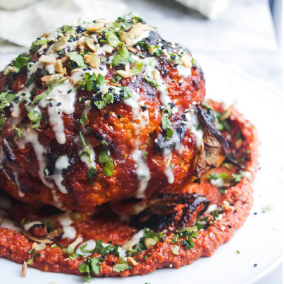 Whole roasted cauliflower on tomato pesto on a white plate, with coriander and sesame seeds on top.