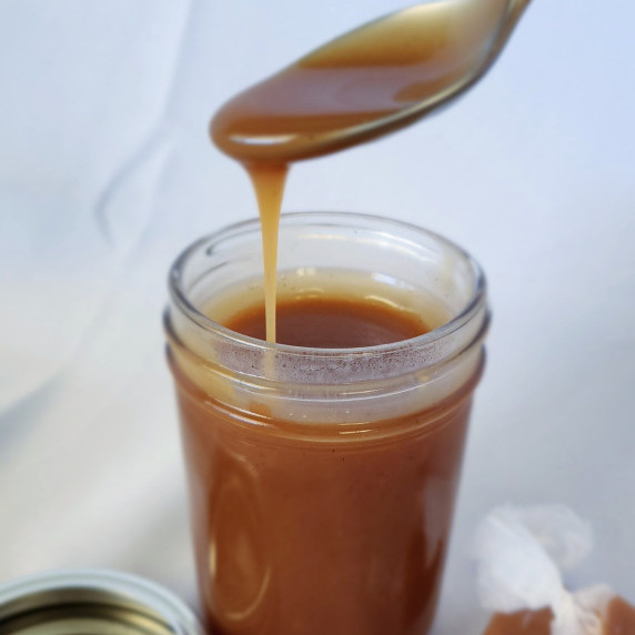 Salted caramel sauce drizzling from a spoon into a jar against a white background.