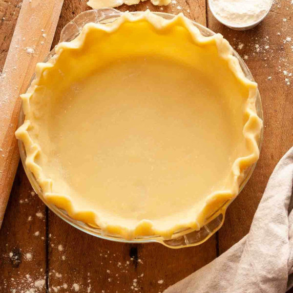 Unbaked pie crust in a glass pie dish on a wooden surface next to a rolling pin and flour.