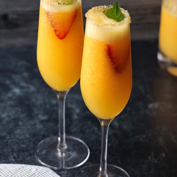 Two rimmed champagne flutes filled with Frozen Peach Bellinis garnished with strawberry and mint.