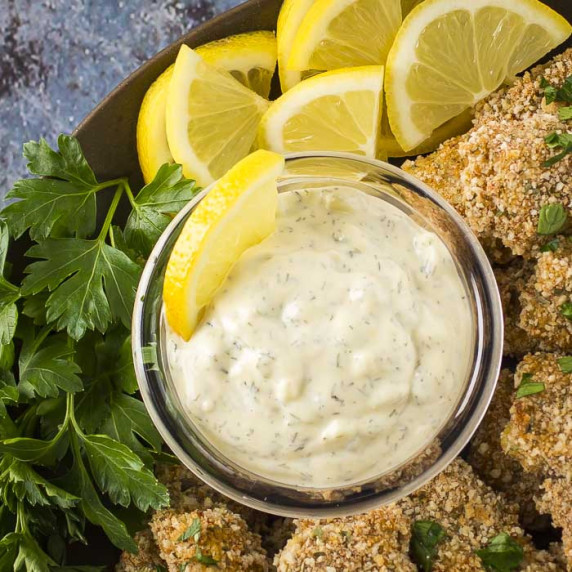 Overhead close up shot of tartar sauce surrounded by breaded fishsticks with lemon wedges and herbs.