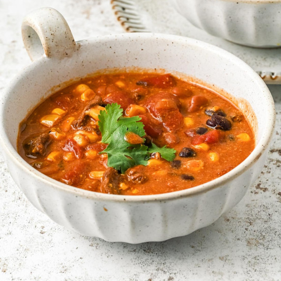 A bowl filled with chili soup containing corn, beans, and chunks of tomato, garnished with cilantro.