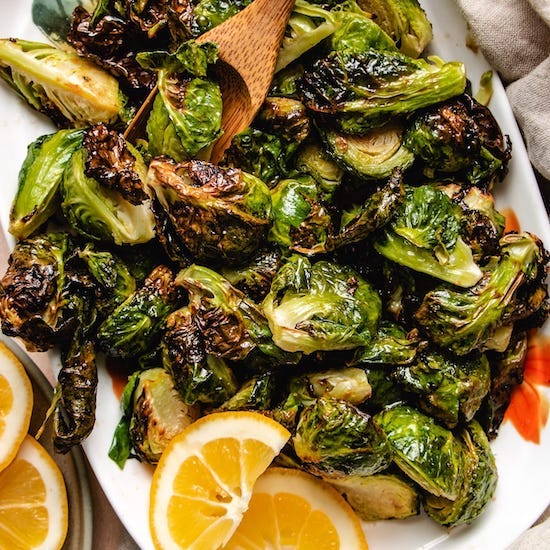 Air fried brussels sprouts with maple glaze and sliced lemons and wooden spoon on a white plate