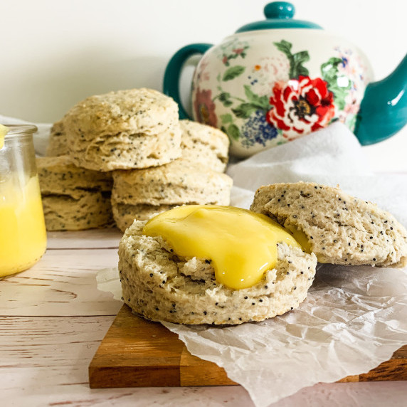 almond poppyseed scone with lemon curd on top beside a jar of curd and a tea pot