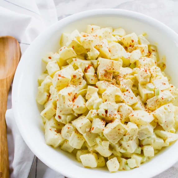 Potato salad garnished with a dust of paprika in a white bowl with a wooden spatula on the side 