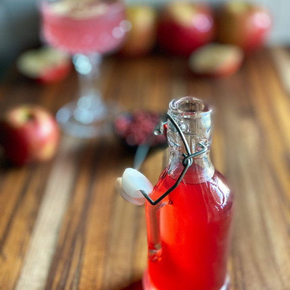 Bottle of pink peppercorn apple simple syrup with apples and peppercorns in background.