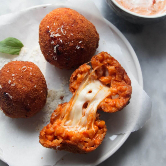 3 arancini balls on a small white plate, one open with mozzarella oozing out, with a bowl of sauce.