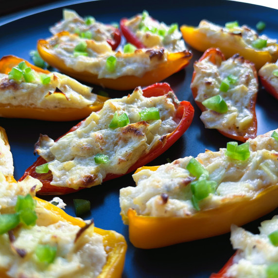 Mini sweet peppers stuffed with an artichoke filling on a black plate topped with diced jalapenos.