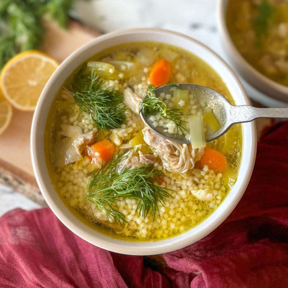 A bowl of chicken pastina soup is shown with a spoon, dill, and lemons.