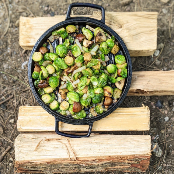 bacon fat brussels with onions and mushrooms in a cast-iron pan on some logs in a campsite