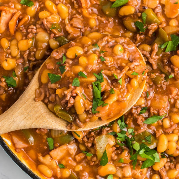 A close-up of a wooden spoon scooping baked beans with ground beef from a pan.