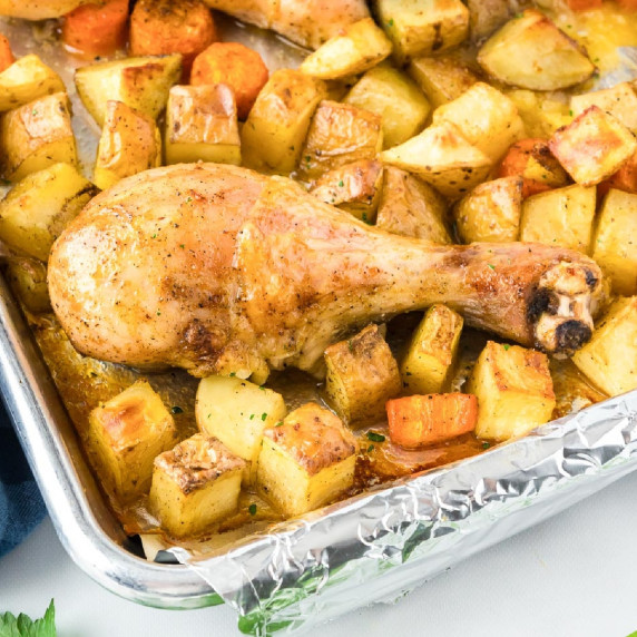 Roasted chicken leg on a bed of golden potatoes and carrots on a foil-lined baking tray.