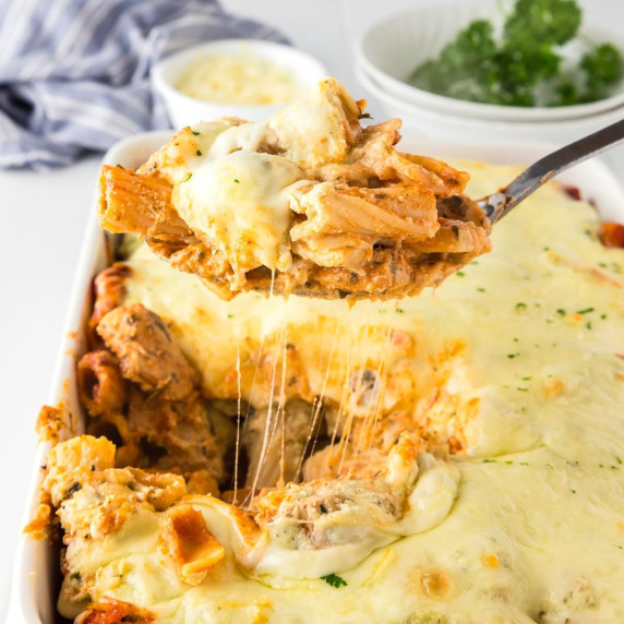 A serving spoon lifts a portion of cheesy baked rigatoni with chicken from a baking dish.