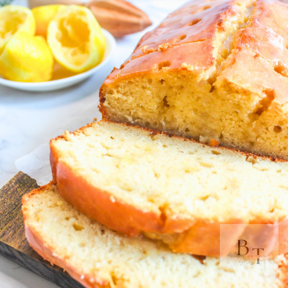 Lemon Loaf on a brown cutting board. Used Lemons in the background in a small white bowl.