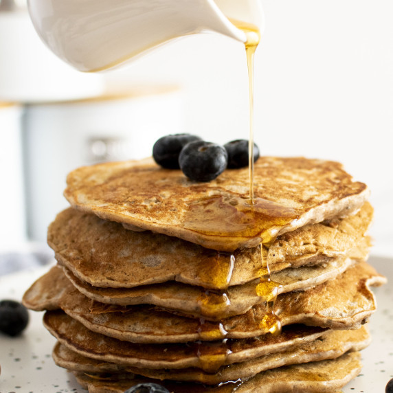 A small jug drizzling maple syrup over a stack of banana buckwheat pancakes.