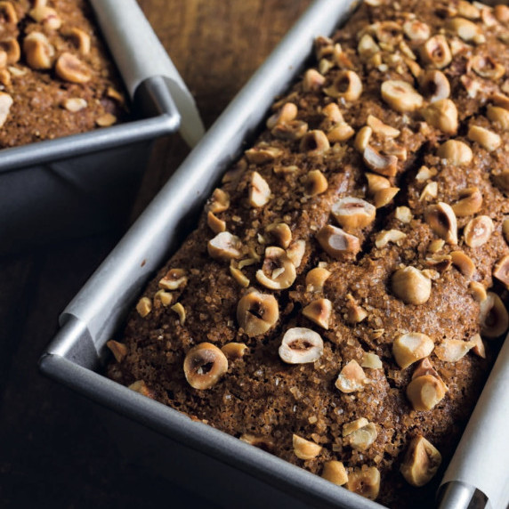 Two loaves of banana hazelnut bread in metal pans, topped with chopped hazelnuts.