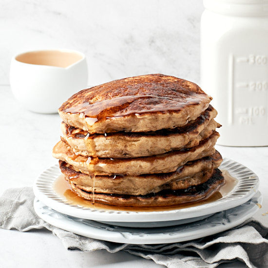 Stack of pancakes on a white plate with maple syrup on a grey napkin on white counter.